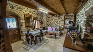A rustic stone-walled room with a wooden ceiling, a dining table with chairs, a sofa, and a small table with a vase of flowers. - Villa Fragk in Nipiditos village (Arkalochori)