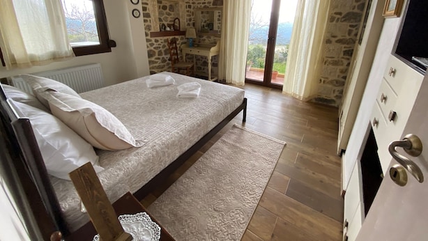 A bedroom with a stone wall, a bed with white linens, a wooden nightstand, a dresser, and a view of the outdoors. - Villa Fragk in Nipiditos village (Arkalochori)