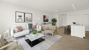 A modern living room with a white sofa, a coffee table, and a kitchen area with a white countertop and bar stools. - 2025 Newly Constructed 3-Bedroom Townhome in Lovely North Fort Myers. (North Fort Myers)