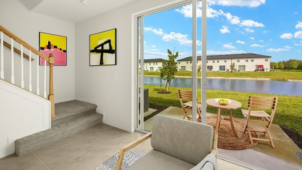 A modern living room with a staircase, a large sliding glass door leading to an outdoor patio with a view of a lake and buildings. - 2025 Newly Constructed 3-Bedroom Townhome in Lovely North Fort Myers. (North Fort Myers)
