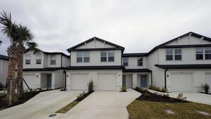 A modern two-story residential building with three attached garages, a concrete driveway, and landscaped areas with small shrubs and a palm tree. - 2025 Newly Constructed 3-Bedroom Townhome in Lovely North Fort Myers. (North Fort Myers)