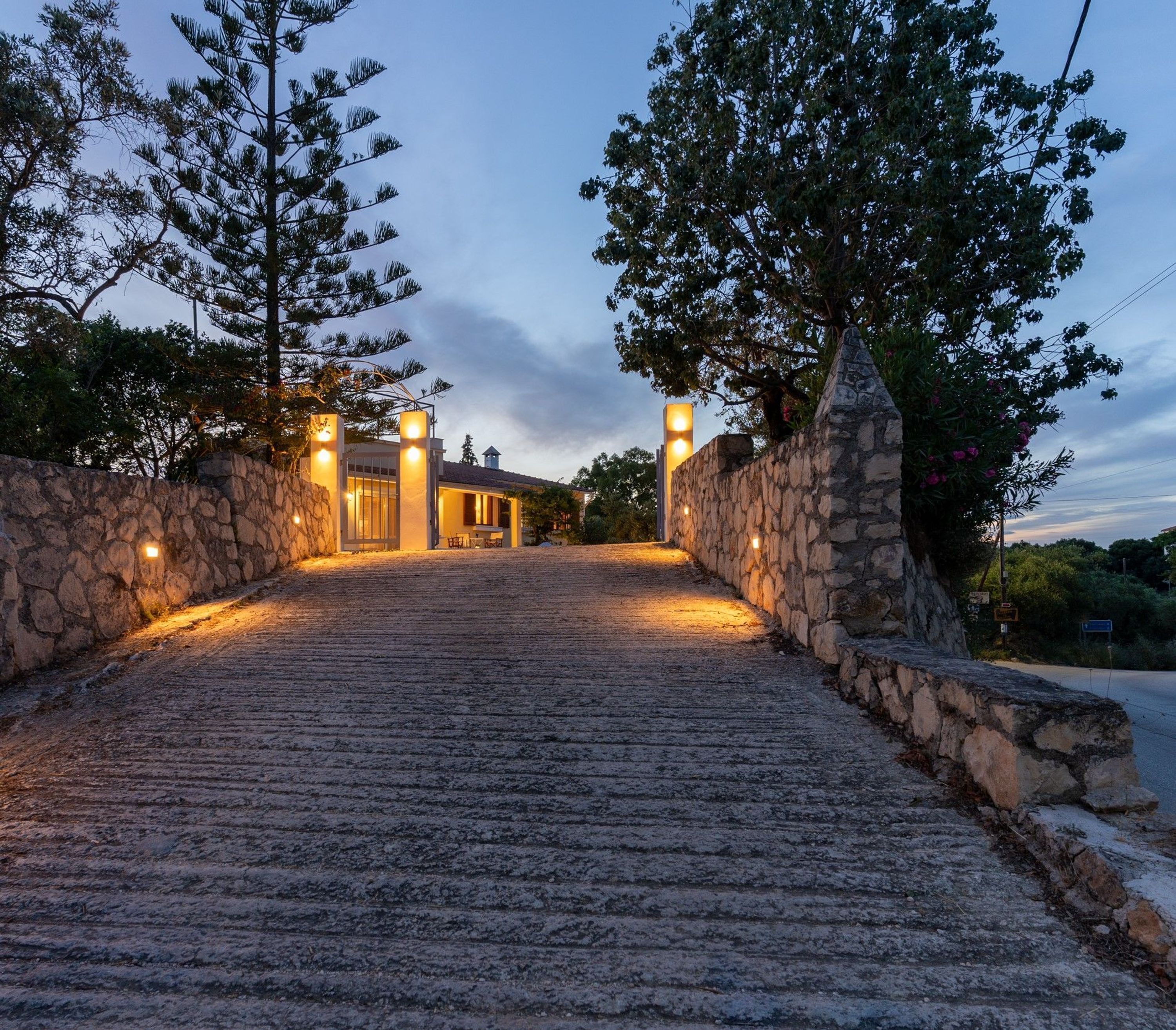 Un chemin de pierres mène à un bâtiment dont un côté est bordé d’un mur de pierres et l’autre de rangées d’arbres.