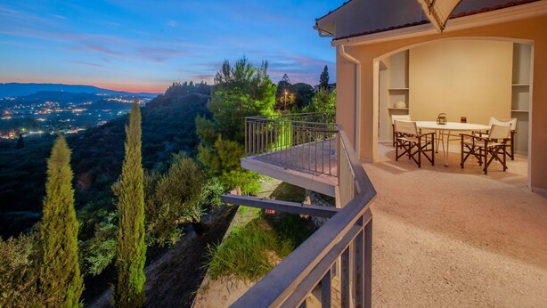 A balcony with a table and chairs, overlooking a scenic landscape with trees and distant lights. - La Belle Villa - Family Retreat & Magnificent View (Zakinthos)