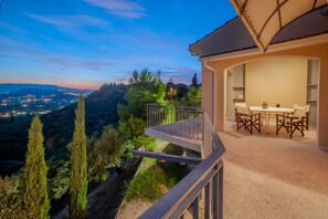 Un balcon avec une table et des chaises, donnant sur un paysage pittoresque, avec des arbres et des lumières scintillantes au loin.