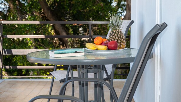 Uma mesa de vidro redonda com uma cadeira de metal, um livro e um prato de frutas, incluindo abacaxi, bananas e maçãs.