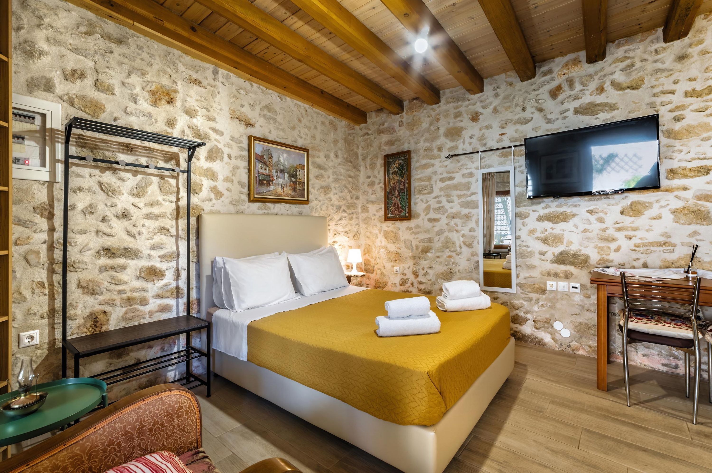 Un dormitorio con pared de piedra, vigas de madera en el techo, una cama con sábanas blancas y una manta color mostaza, un televisor montado en la pared, y una pequeña mesa con una silla.