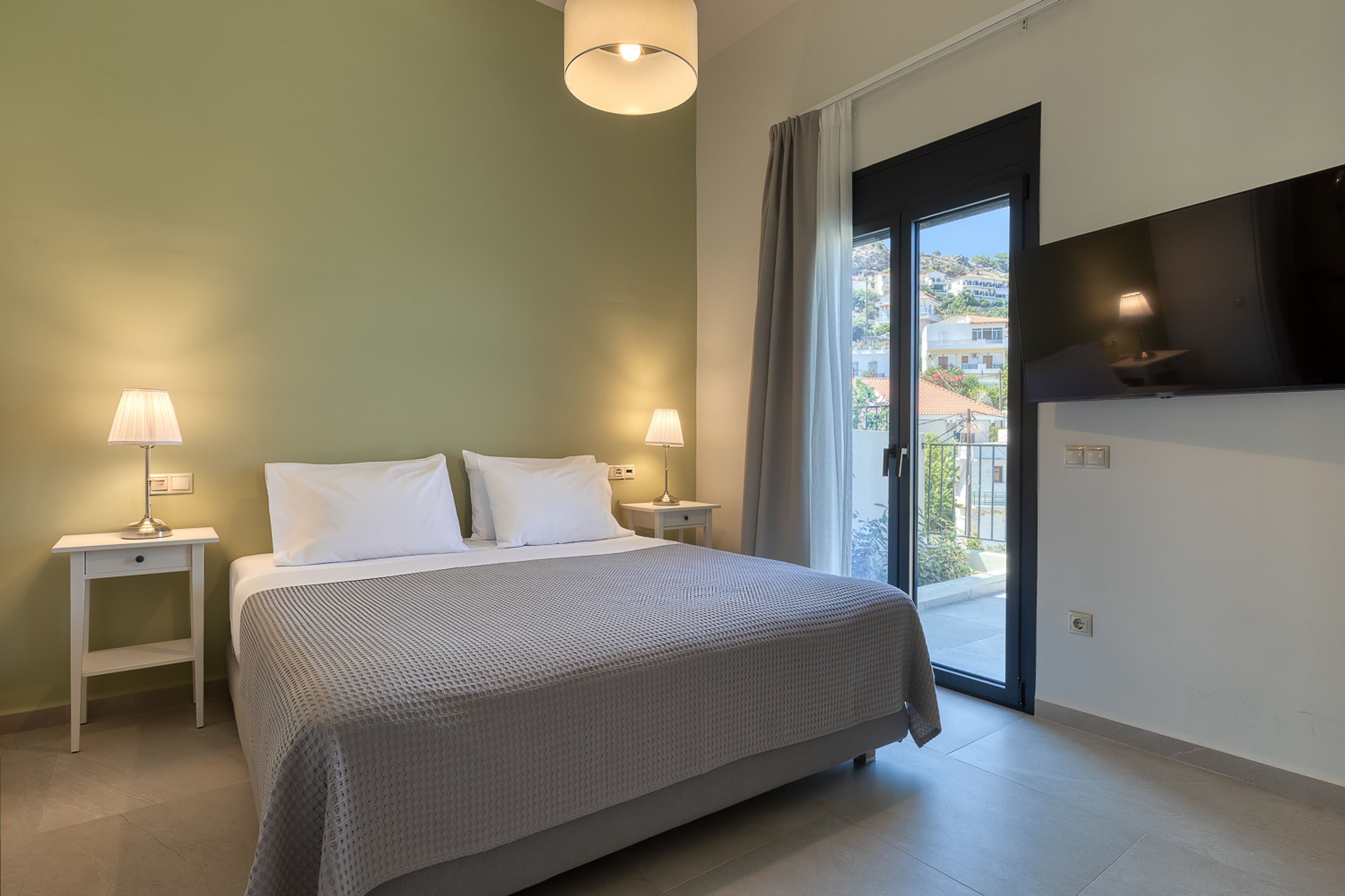 A bedroom with a bed, two bedside tables with lamps, a TV mounted on the wall, and a sliding glass door leading to an outdoor area.