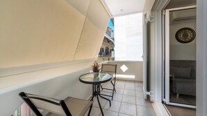 En balkon med et bord og stole, en skydedør til et rum, og udsigt til bygninger i baggrunden.