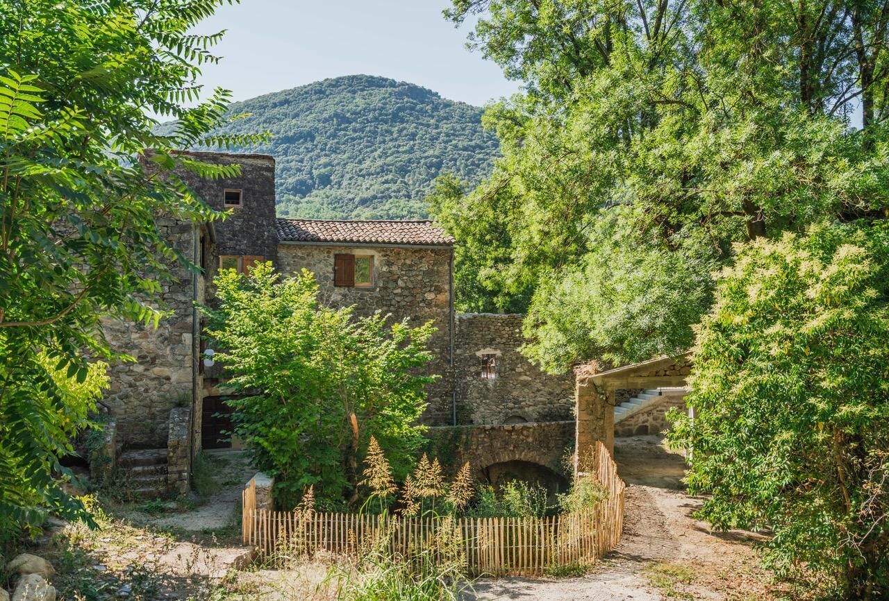 Un edificio rústico de piedra con techo de tejas, rodeado de exuberante vegetación y una montaña al fondo.