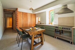 A modern kitchen with a wooden dining table, green chairs, a stainless steel oven, and a window with a view of greenery. - The mill in the Cévennes (Saint-Jean-du-Gard)