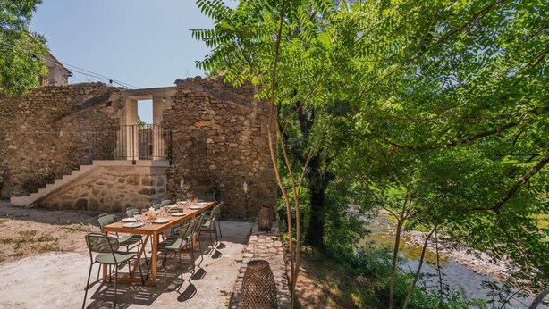 Un edificio de piedra con una mesa y sillas de madera instaladas al aire libre, rodeado de follaje y un arroyo.