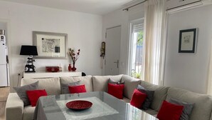 Una sala moderna con una mesa de centro de vidrio, un sofá color beige con cojines rojos y grises, una mesa de comedor blanca y una cocina al fondo.