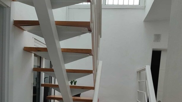 Una escalera blanca con escalones de madera y barandillas blancas en una habitación con paredes blancas y una ventana.