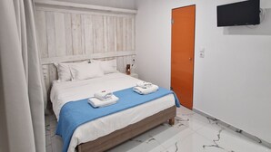 Une chambre à coucher avec un lit, des rideaux blancs, une tête de lit en bois, une couverture bleue, un téléviseur fixé au mur et un sol à motifs de marbre.