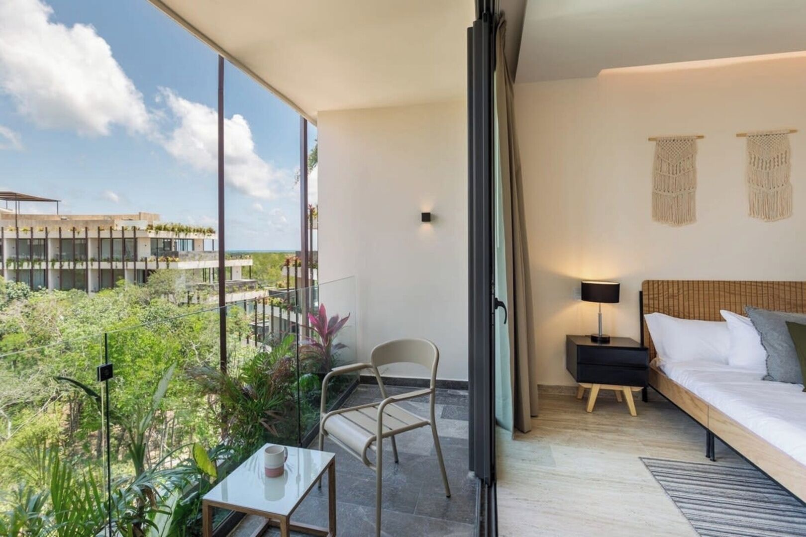 En balkon med et bord og en stol, en seng med et natbord, og udsigt til en bygning og grønt.