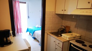Une cuisine compacte équipée d’une cuisinière, d’un évier et d’un petit plan de travail. On aperçoit un lit recouvert d’une courtepointe bleue et blanche à l’arrière-plan.