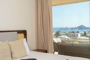 A hotel room with a bed, a nightstand, and a view of the sea and buildings. - Oceanview Escape • Fast Wi-Fi • AC • Chic Design (Cabo San Lucas)