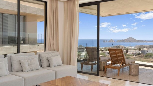 A modern living room with a sofa, a coffee table, and two wooden chairs. Large glass doors lead to a balcony with a view of the sea and buildings. - Enjoy your perfect vacation in this 3 bd (Cabo San Lucas)