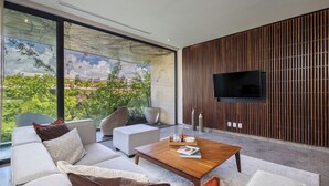 A modern living room with a large window, a flat-screen TV mounted on a wooden panel wall, and a comfortable seating area with sofas and a coffee table. - Chic 3BR w/ Private Pool, Gym and Stunning Views (Tulum)