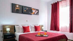 Een hotelkamer met een bed, rode gordijnen, een nachtkastje, een kunstwerk aan de muur en een dienblad met jus d'orange en gebak.
