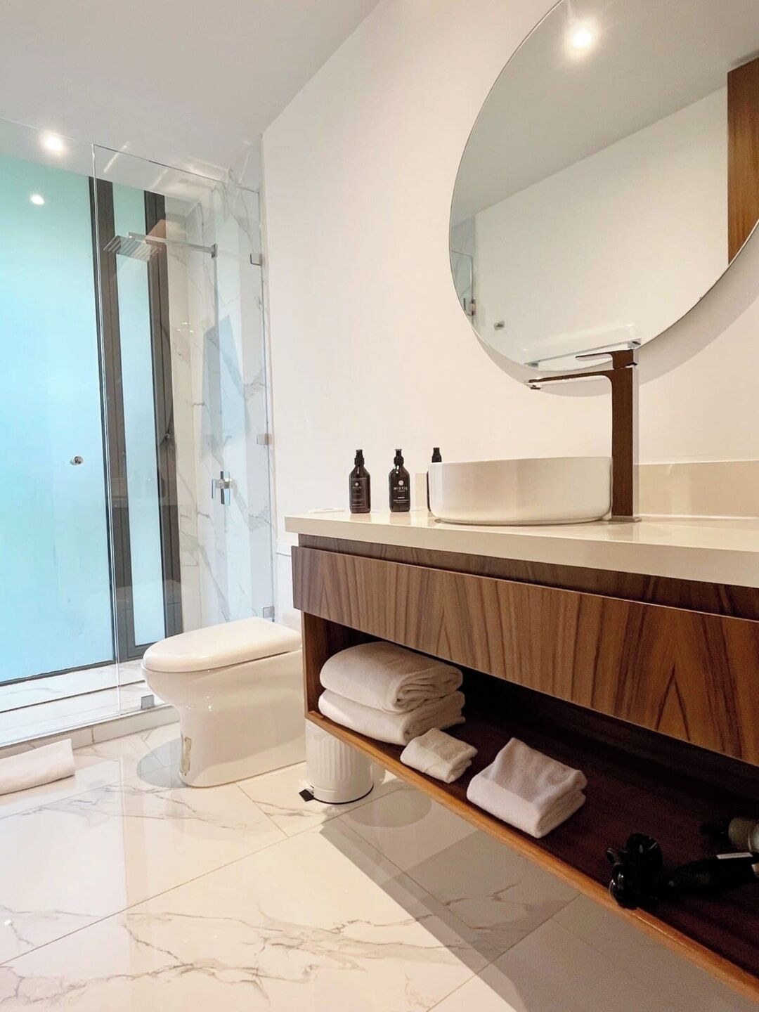 Une salle de bain moderne avec une douche à parois de verre, des toilettes, un meuble-lavabo avec une étagère en bois et un grand miroir.