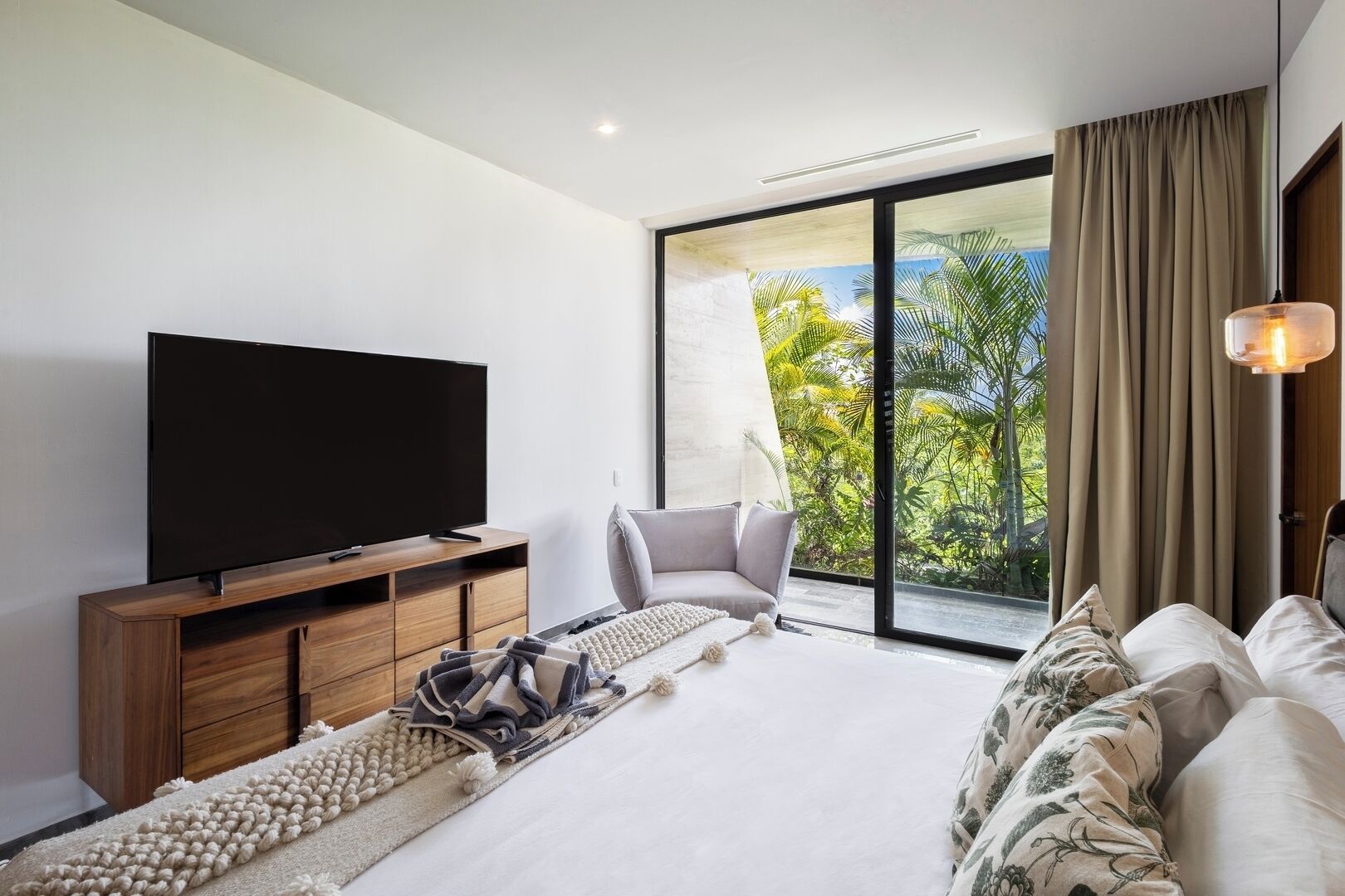 Une chambre moderne avec un téléviseur à écran plat posé sur un meuble en bois, un grand lit avec une literie à motifs, un canapé gris et une vue sur une végétation luxuriante, visible à travers une porte-fenêtre coulissante.
