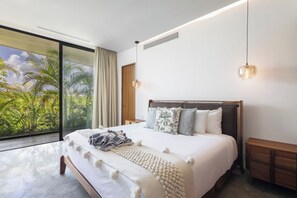 Une chambre moderne avec un grand lit, une table de chevet, une tête de lit en bois et une vue sur les palmiers, visible à travers une porte-fenêtre coulissante.