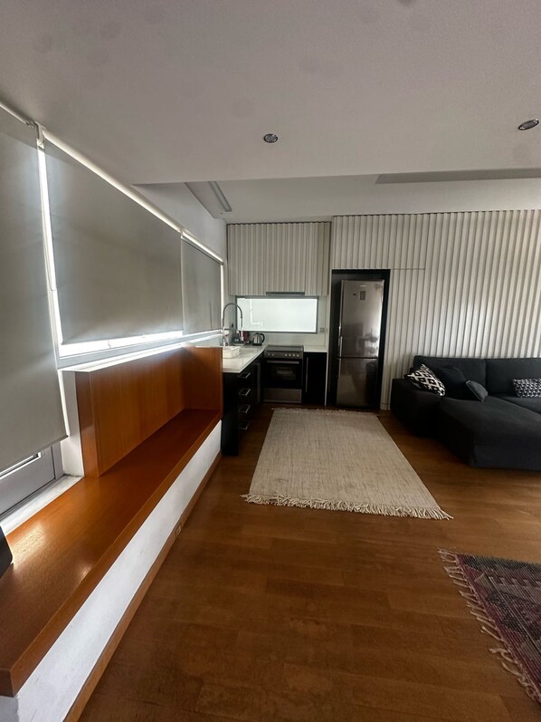 Un salon moderne avec un banc en bois, un canapé noir et un coin cuisine équipé d’un réfrigérateur et d’une cuisinière.