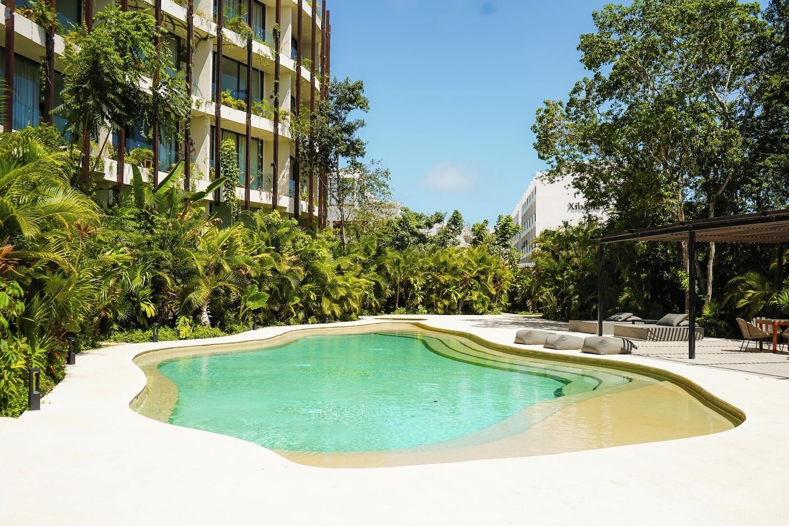 A swimming pool with a curved edge, surrounded by lush greenery and a building with balconies.
