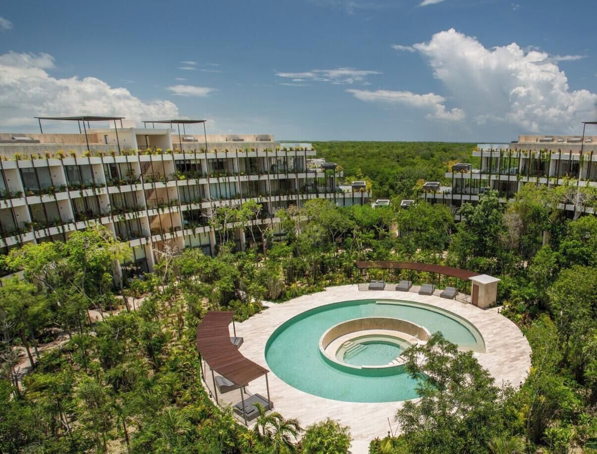 A modern hotel with a circular pool surrounded by lush greenery and a clear blue sky.