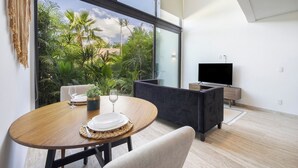 Uma sala de estar moderna com uma janela ampla, uma mesa de madeira redonda com pratos e um copo, um sofá escuro e uma televisão de tela plana em um suporte de madeira.