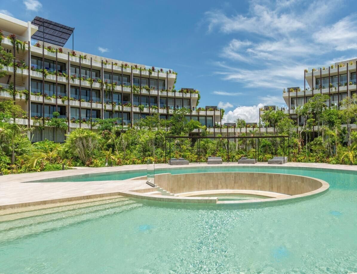 A modern hotel with a large pool, lush greenery, and a clear blue sky.