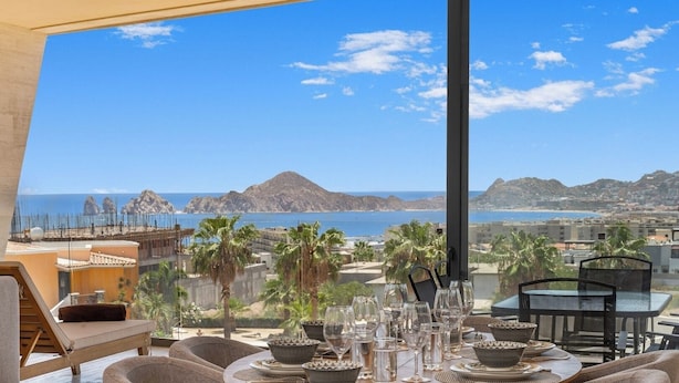 A dining area with a table set for a meal, overlooking a coastal landscape with mountains and the sea. - Bright apartment w/ ocean view and unique decoration (Cabo San Lucas)