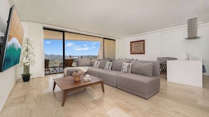A spacious living room with a large grey sofa, a wooden coffee table, and a view of the city through large windows. - Bright apartment w/ ocean view and unique decoration (Cabo San Lucas)