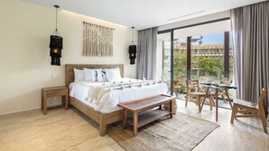 Une chambre dotée d’un grand lit, de tables de chevet en bois, d’un banc, d’un coin repas avec des chaises et offrant une vue sur la verdure grâce à de grandes fenêtres.