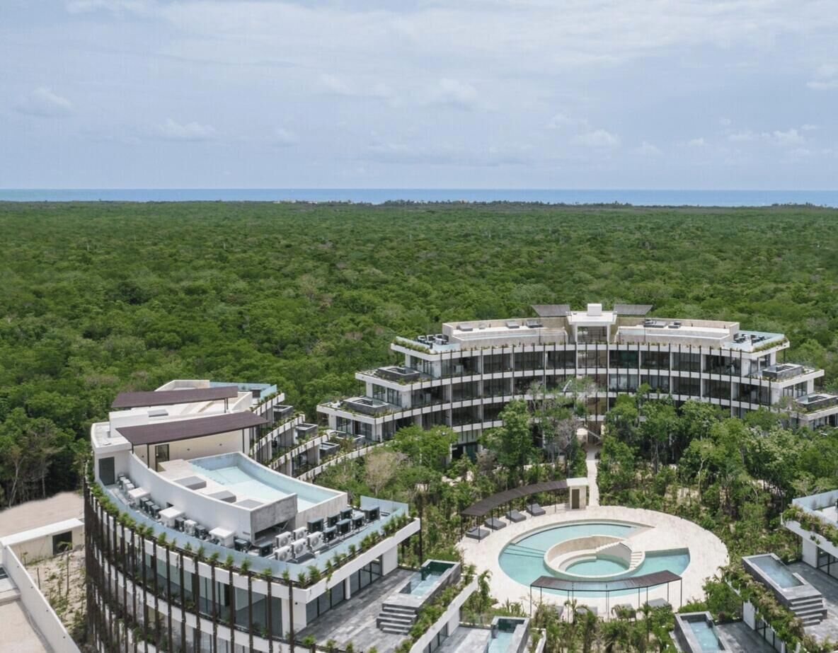 A modern hotel with a circular pool, surrounded by lush greenery and a clear view of the ocean.