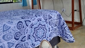A room with a bed, a TV on a wooden stand, a window with blue curtains, and a patterned bedspread. - M&K House - Room MK Double first level (Tarapoto)