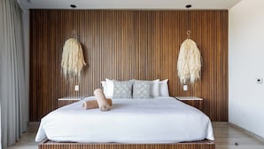 A modern bedroom with a large bed, wooden panel wall, two hanging light fixtures, and a bedside table. - Lavish 3 BD Apartment with Private Pool (Tulum)
