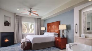 Una camera d'albergo con un letto, un camino, una ventola a soffitto, un comodino con una lampada, un bagno con una vasca e un grande specchio.