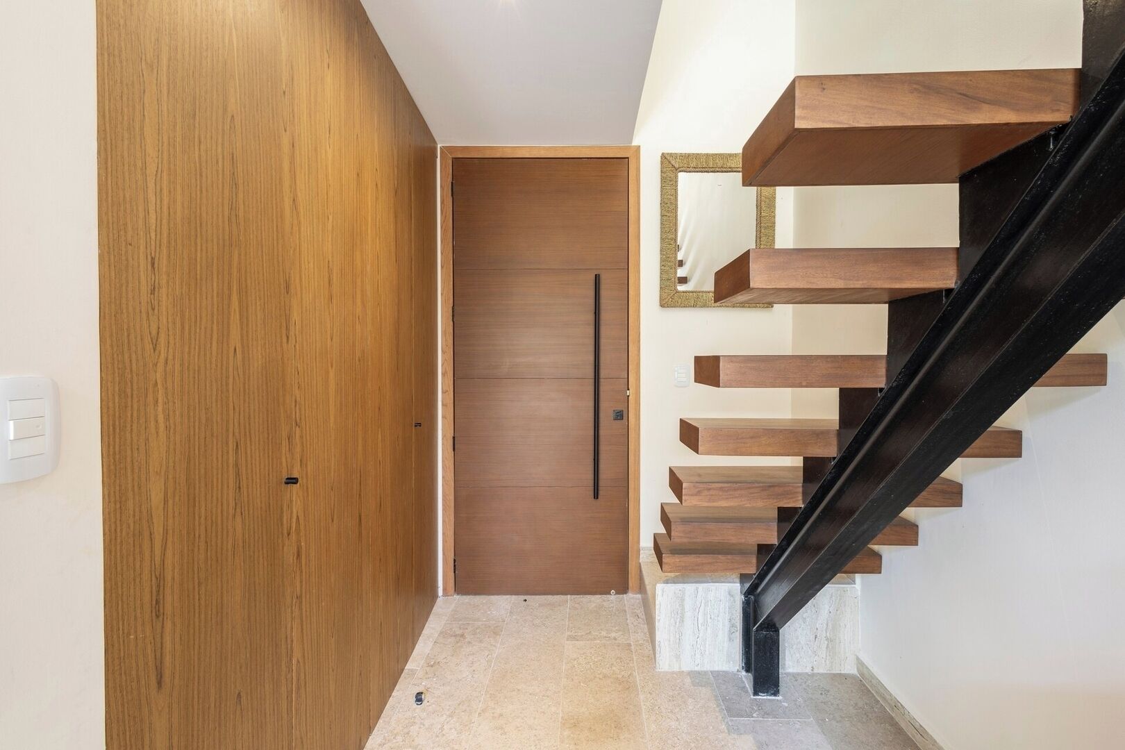 A modern interior with a wooden door, a staircase with wooden steps and a black handrail, and a light-colored floor.