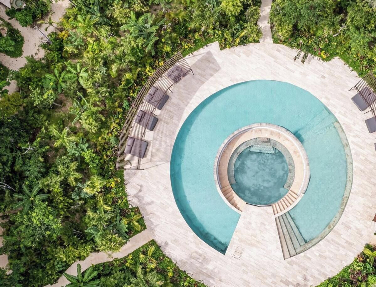 Vista aérea de una piscina infinita circular con un jacuzzi central, rodeada por una terraza de piedra y exuberante vegetación tropical.