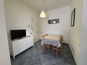 A small, clean room with a TV on a white cabinet, a dining table with chairs, and a hanging light fixture. - Apartment in a building facing the sea-Rif. Giglio (San Vincenzo)