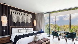 A modern bedroom with a large bed, a bench, a dining area with chairs, and a view of trees through large windows. - Stylish Studio with gym, pool (Tulum)