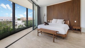 Une chambre moderne avec un grand lit, un banc en bois, une table de chevet et une vue sur les bâtiments à travers de grandes fenêtres.