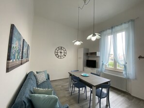 A modern living room with a sofa, dining table, and chairs. There is a clock on the wall and a window with curtains. - 5 bed places with parking space - ROSS2S-Rif. Ross2S (San Vincenzo)
