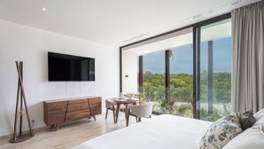 A modern bedroom with a large bed, a TV mounted on the wall, a wooden dresser, and a dining area with a view of the outdoors. - Jungle view studio, AC, Wifi , Several Pools (Tulum)