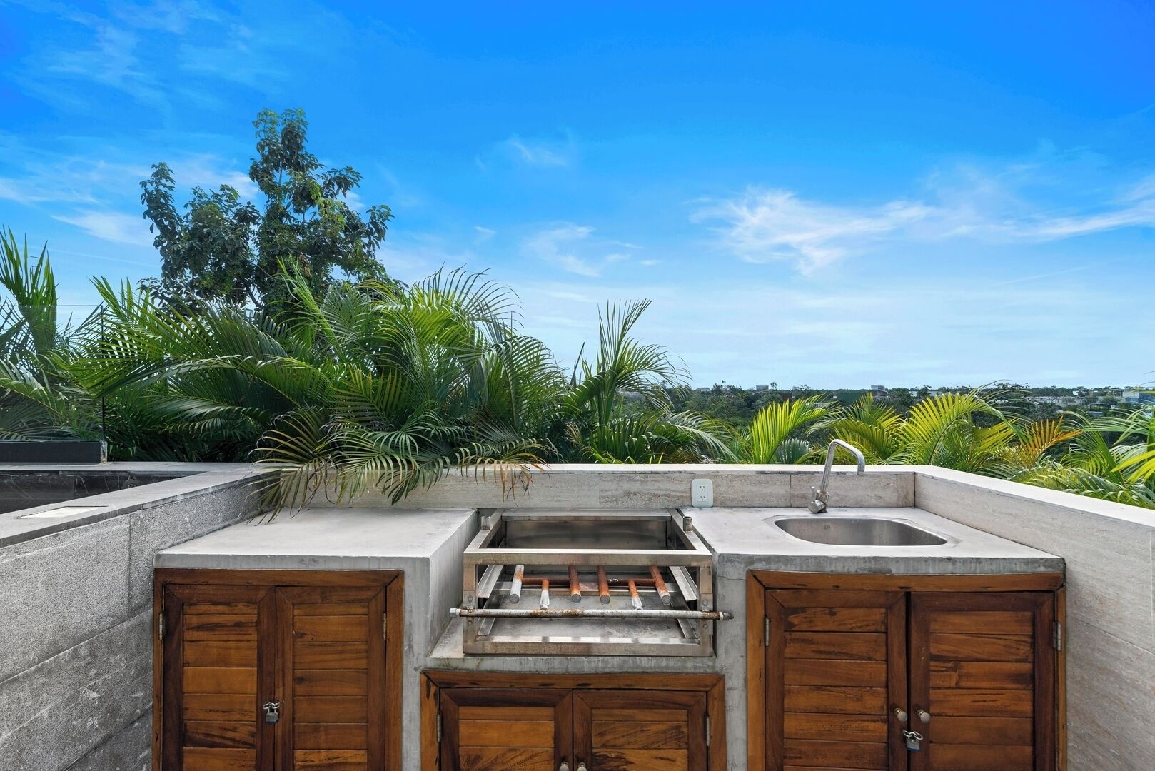 An outdoor kitchen with a sink and grill, surrounded by lush greenery and a clear blue sky.