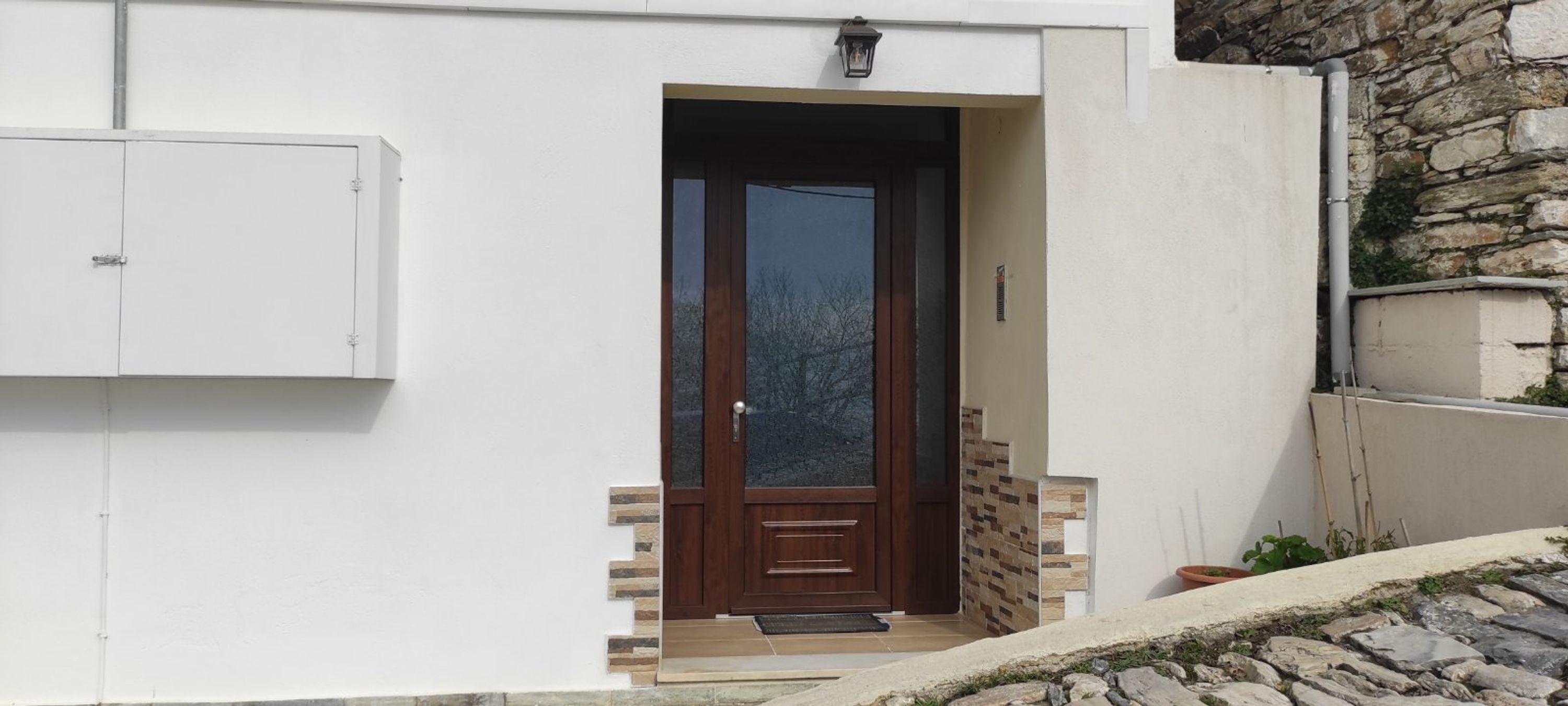 A white building with a wooden door, a small set of steps leading to the entrance, and a stone wall to the right.
