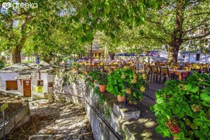 Zona esterna con tavoli e sedie all'ombra degli alberi, un muro in pietra e un piccolo edificio con porta e finestra.