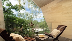 A modern outdoor seating area with two wooden lounge chairs and a woven basket on a small table, overlooking a lush garden and a clear sky. - Exclusive 2 bd with pool and street view! WiFi and AC (Tulum)